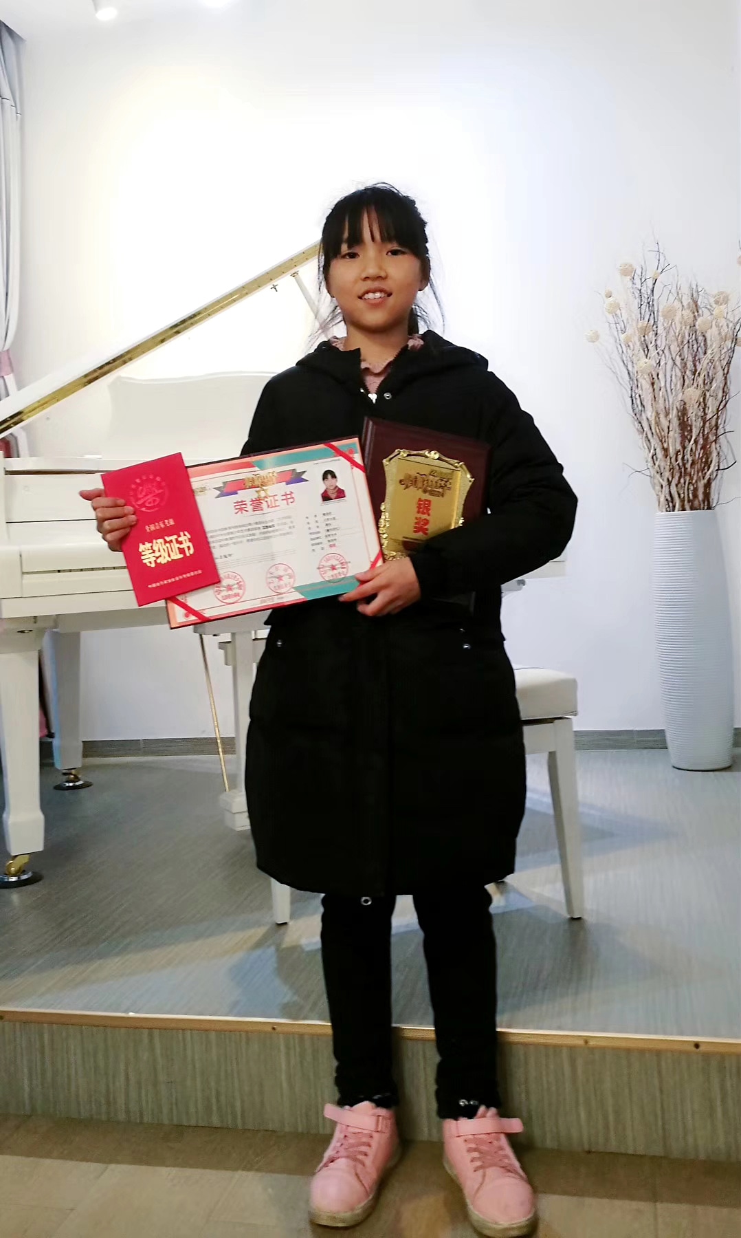 学生站在白色三角钢琴前手持证书、考级证书与奖牌